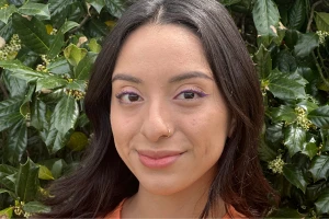 Yesenia with long dark hair and nose ring smiling near green bushes